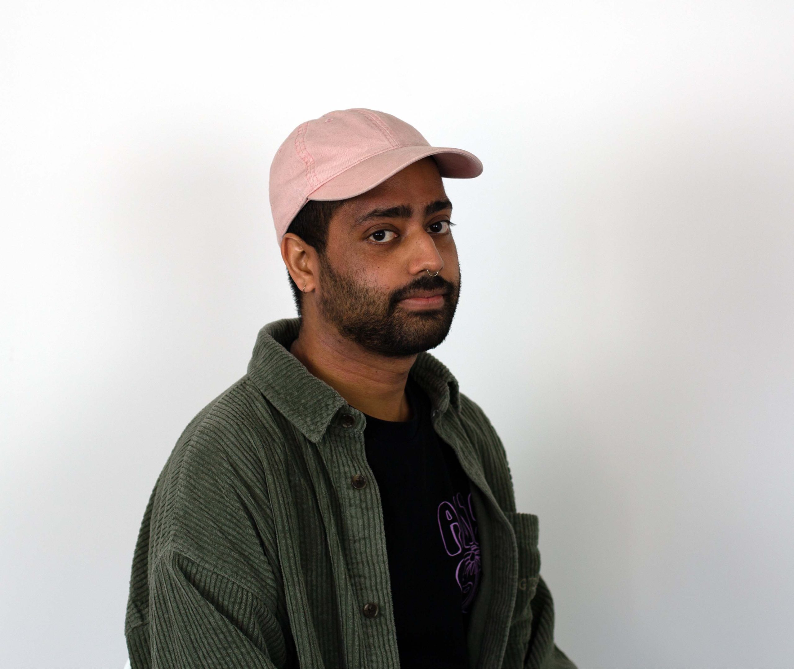 A headshot of Arjun, a Indian man with black stubble, a golden nose ring and pink hat wearing a green corduroy shirt over a.black t-shirt.