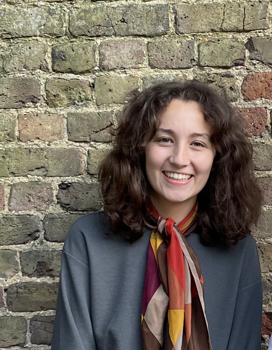 A headshot of Kaiya who has long brown curly hair, and is wearing a red, brown and yellow neckerchief and teal jumper. She is also standing against a brown brick wall.